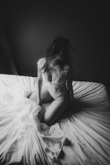 Schwarz-Weiß-Boudoiraufnahme einer Frau in weißer Spitze auf einem Bett mit weich fallenden Stoffen.