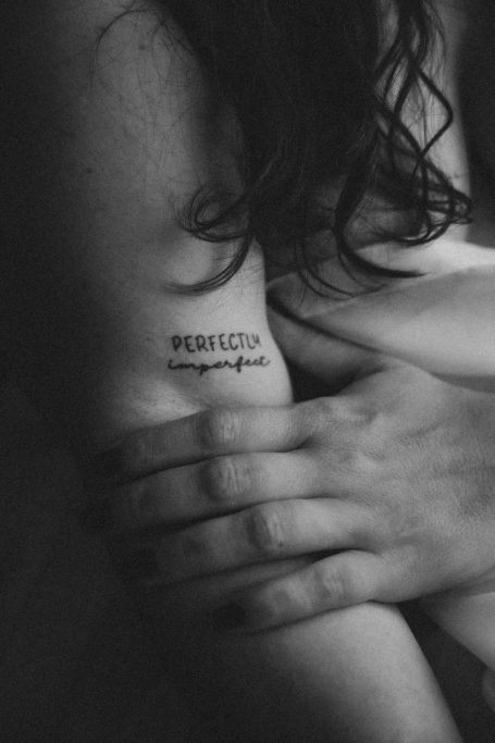 Schwarzweiß-Porträt einer Frau mit dunklem Haar, die sich mit den Händen umarmt. Auf ihrer Haut steht „Perfectly imperfect“.