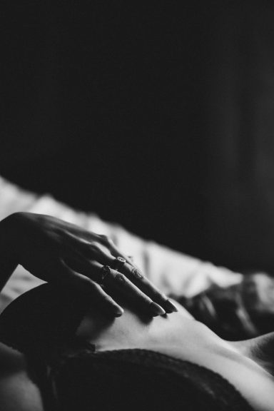 Intime Schwarz-Weiß-Detailaufnahme einer Hand mit Ringen auf dem Oberkörper einer Frau in schwarzer Spitze.