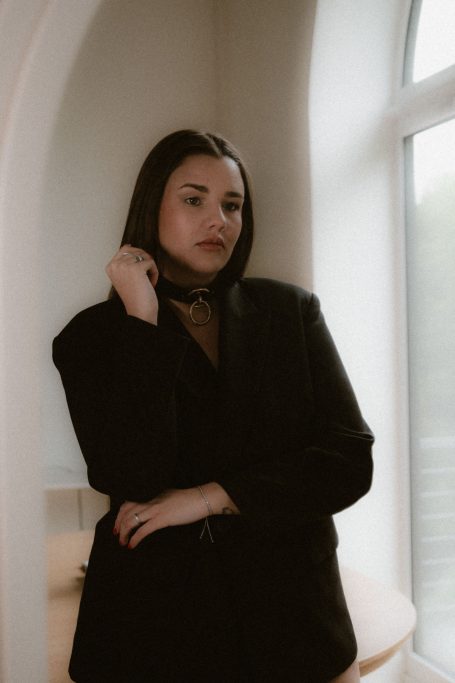 Elegantes Porträt einer Frau im schwarzen Blazer mit auffälligem Halsband neben einem hellen Fenster.