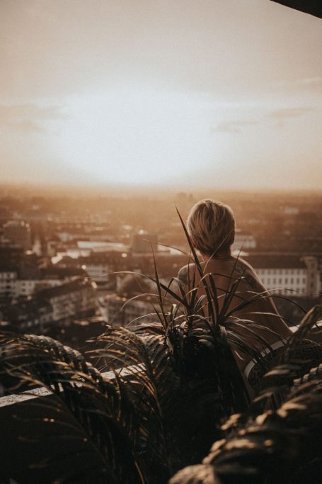 Nackte Frau mit dem Rücken zur Kamera auf einem Dach über einer Stadt im warmen Abendlicht.