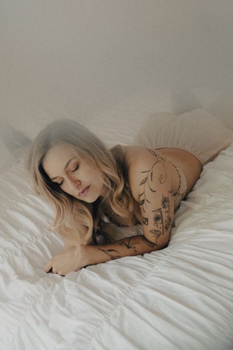 Frau liegt in heller Boudoir-Lingerie auf einem weißen Bett und blickt mit geschlossenen Augen zur Seite