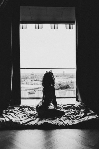 Silhouette einer sitzenden Frau auf einem Bett vor einem großen Fenster mit weitem Stadtblick.
