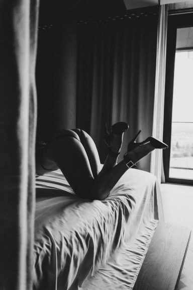 Schwarz-Weiß-Aufnahme einer Frau mit angewinkelten Beinen und High Heels auf einem Bett am Fenster.