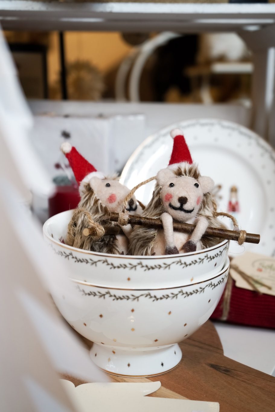 Zwei kleine Filz-Igel mit roten Weihnachtsmützen in weißen Schalen mit goldenen Details.
