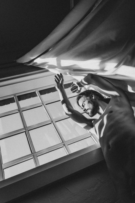 Unbekleideter Mann vor einem großen Fenster, fotografiert aus tiefer Perspektive mit grafischem Licht- und Schattenspiel