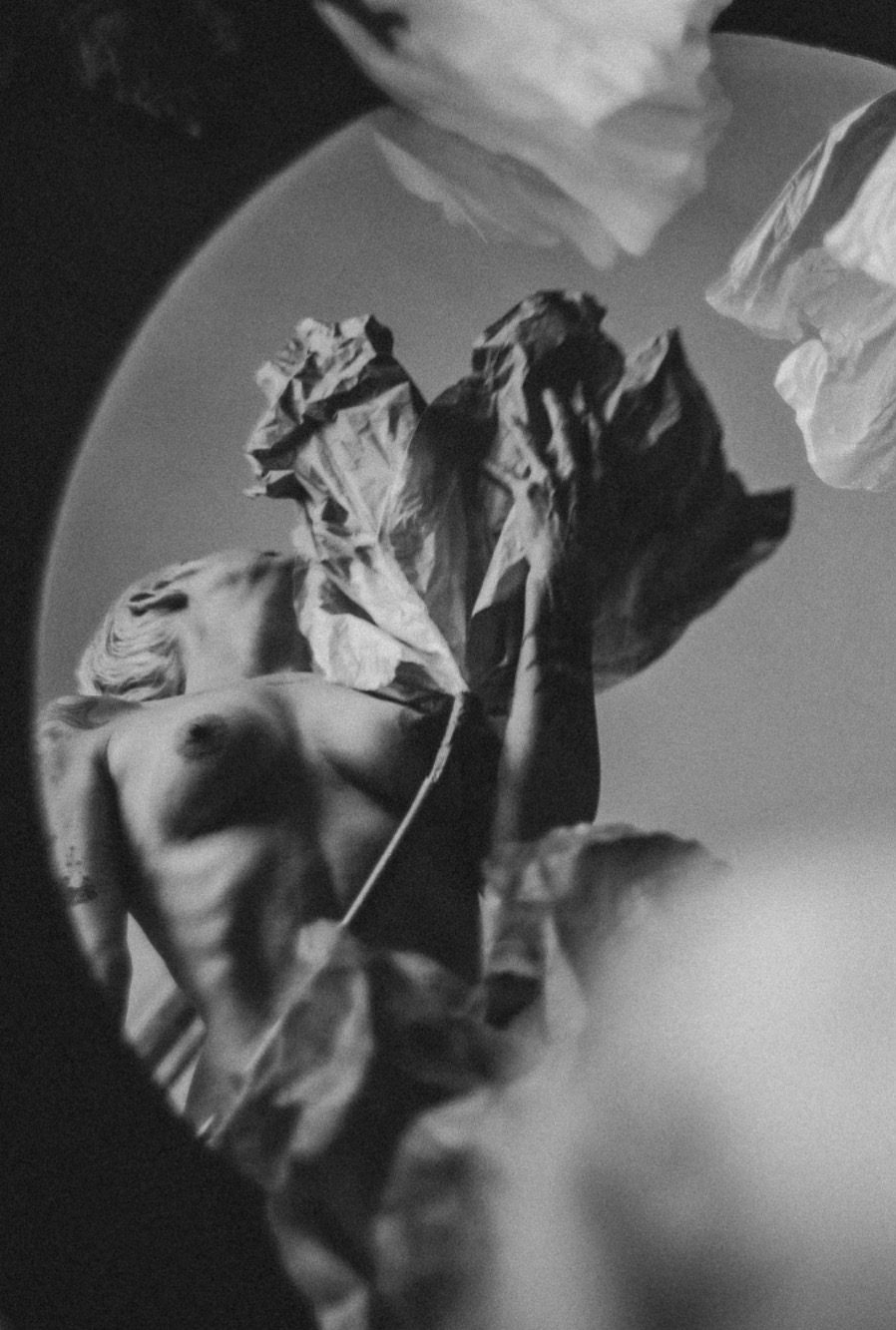Künstlerische Schwarz-Weiß-Aktaufnahme einer Frau, die zwischen großen Papierblumen in einem runden Spiegel reflektiert wird