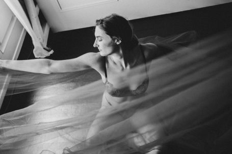 Schwarz-Weiß-Boudoiraufnahme einer Frau mit ausgestrecktem Arm und fließendem transparentem Stoff im Vordergrund.