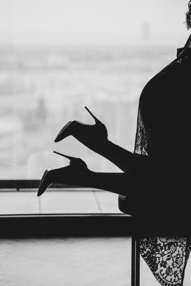 Schwarz-Weiß-Silhouette einer Frau in Spitzenwäsche und High Heels vor einem hellen Fenster.