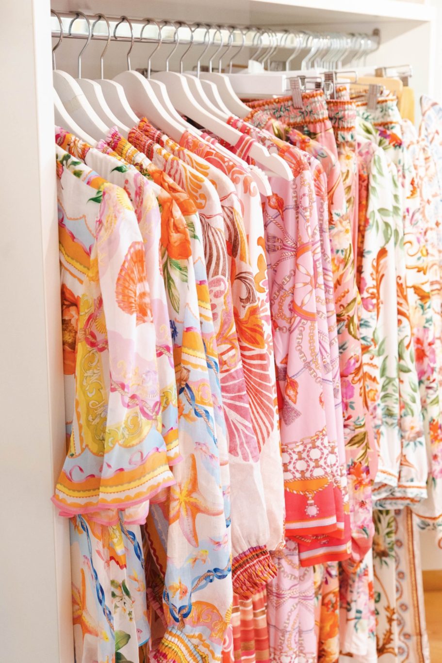 Bunte Sommerkleidung mit floralen und grafischen Mustern in Rosa, Orange und Grün auf weißen Bügeln.