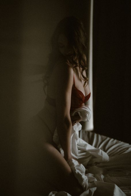 Frau in roter Lingerie hält weiße Bettwäsche vor dem Körper in einer dunkel inszenierten Boudoiraufnahme.