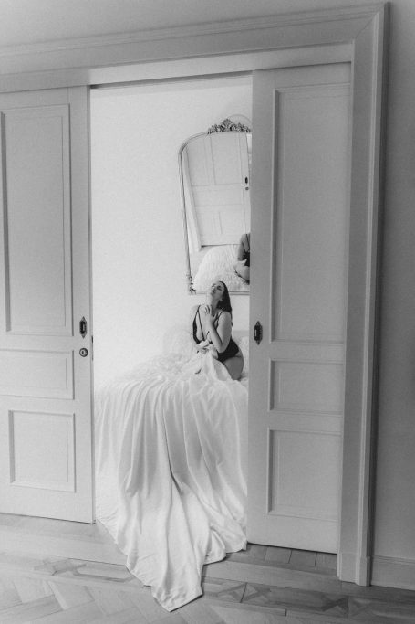 Schwarz-weißes Boudoir-Porträt einer Frau in schwarzer Lingerie, die auf einem weißen Bett vor einem antiken Spiegel sitzt