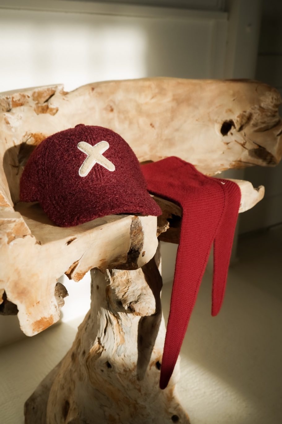Bordeauxrote Cap mit hellem Kreuzdetail und passender Strickschal auf einem rustikalen Holzstuhl.