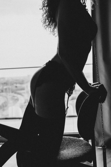 Kontrastreiche Schwarz-Weiß-Silhouette einer Frau in Lingerie und Strümpfen, die an einem Stuhl vor dem Fenster kniet.