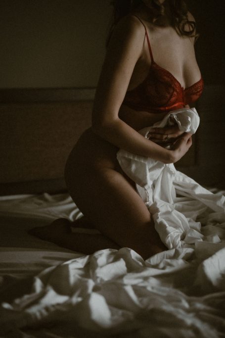 Frau in roter Spitzenwäsche kniet auf einem Bett und hält weiße Bettwäsche in den Händen.