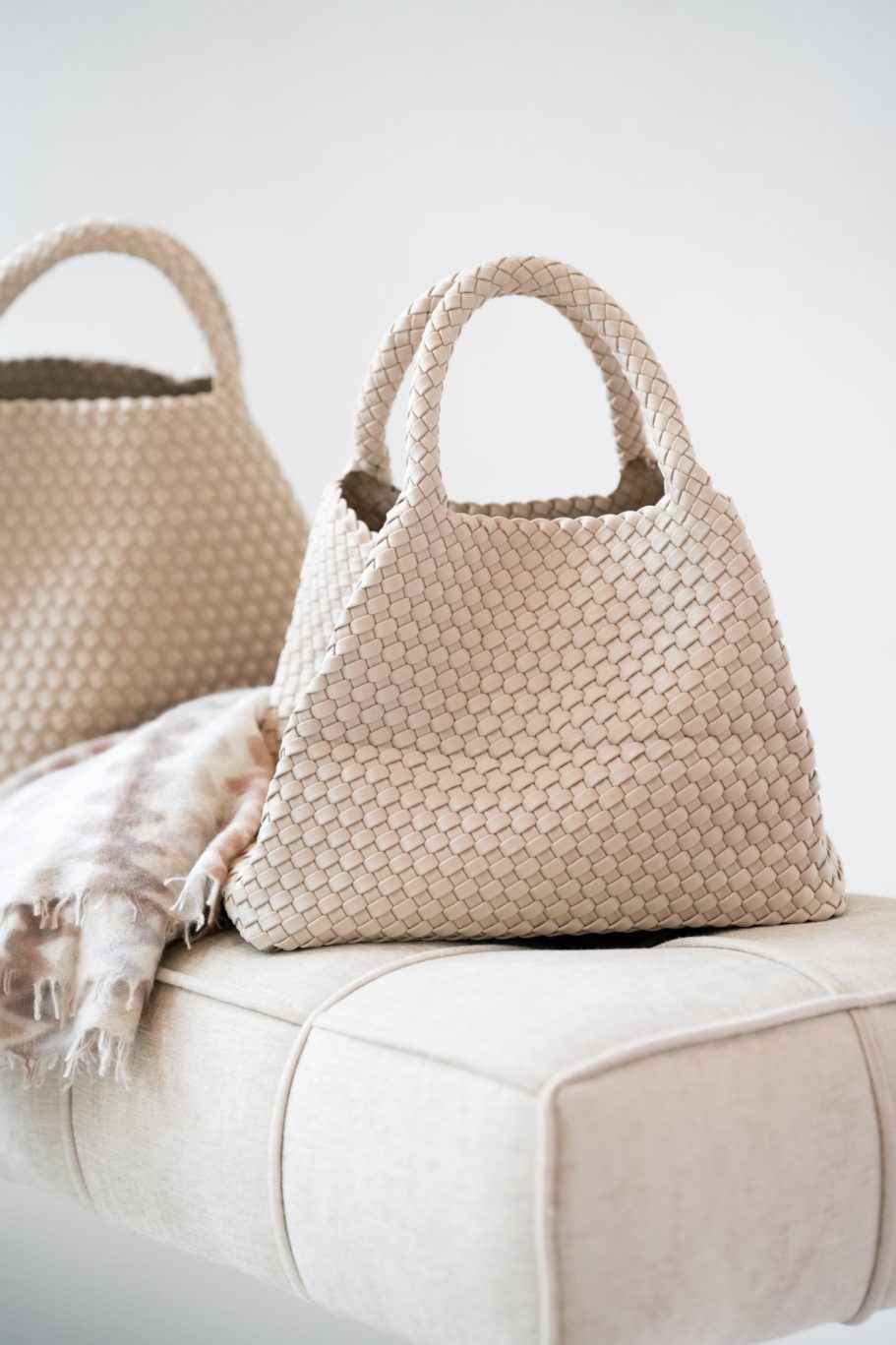 Zwei beige geflochtene Handtaschen auf einem hellen Polster mit weichem, natürlichem Licht.