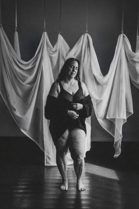Schwarz-Weiß-Boudoir-Porträt einer kurvigen Frau mit Lipödem in schwarzer Lingerie und Morgenmantel vor hängenden Stoffbahnen.