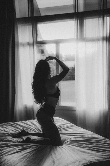 Künstlerische Schwarz-Weiß-Boudoiraufnahme einer Frau im schwarzen Body, die auf einem Bett vor einem großen Fenster posiert.
