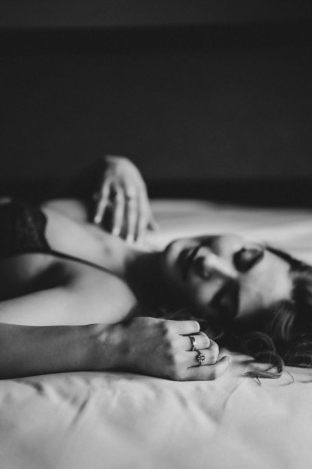 Schwarz-Weiß-Boudoiraufnahme einer Frau, die entspannt auf einem Bett liegt, mit ihren Händen und Ringen im Vordergrund.