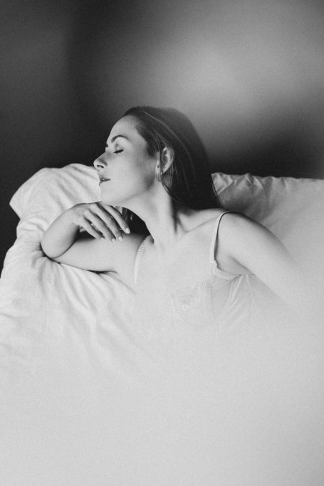Frau liegt entspannt in heller Lingerie auf einem Bett, fotografiert in sanftem Schwarz-Weiß.