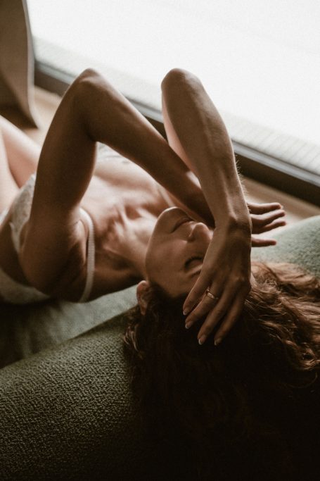 Frau in weißer Lingerie liegt entspannt auf einem Sofa am Fenster und hält die Hände vor ihr Gesicht.