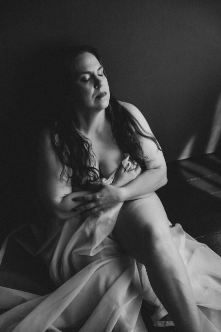 Schwarz-Weiß-Boudoiraufnahme einer Frau, die sich mit geschlossenen Augen selbst umarmt und von weichem Stoff umgeben ist.