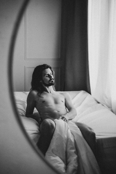 Unbekleideter Mann mit langen Haaren sitzt auf einem Bett und wird über einen Spiegel in Schwarz-Weiß fotografiert