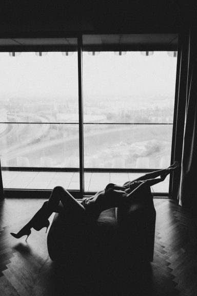 Frau liegt in High Heels auf einem Sessel vor einem großen Fenster, als kontrastreiche Schwarz-Weiß-Silhouette fotografiert.
