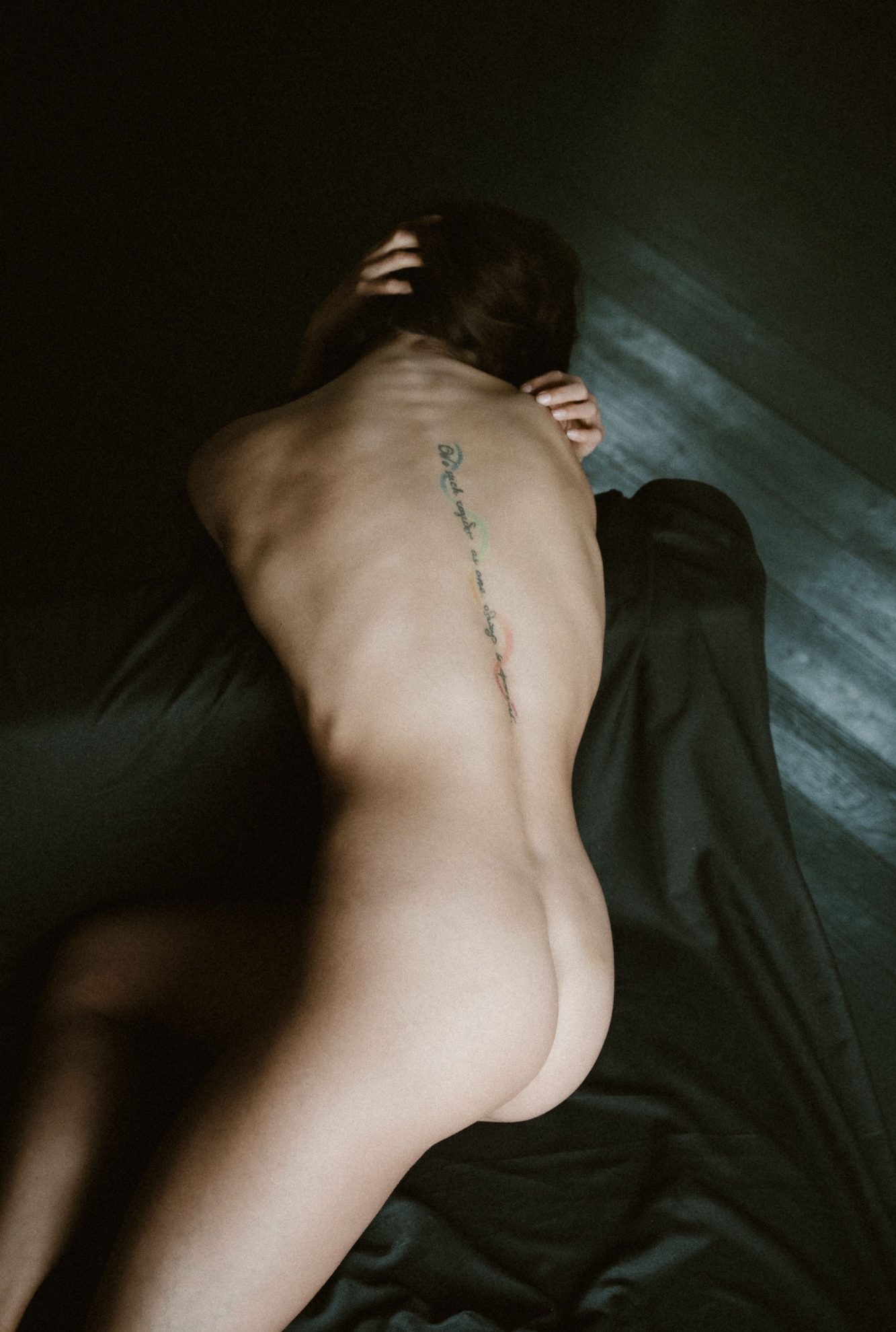 Fine-Art-Aktaufnahme einer unbekleideten Frau von hinten mit sichtbarem Rückentattoo auf dunklem Stoff