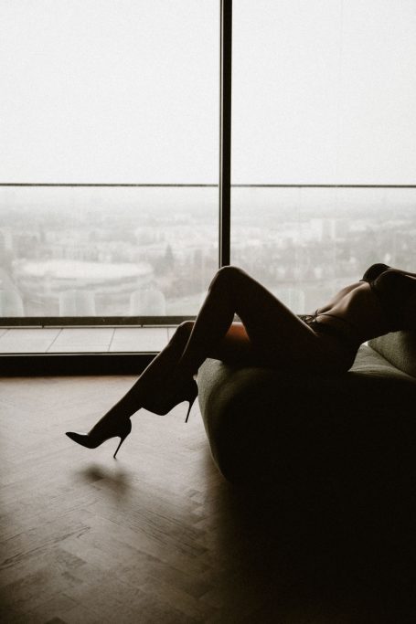 Silhouette einer Frau in High Heels, die entspannt vor einem großen Fenster auf einem Sessel liegt.