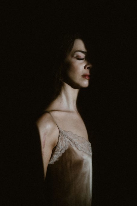 Stimmungvolles Boudoir-Porträt einer Frau in einem Satin-Top mit Spitze, teilweise von tiefem Schatten umgeben.