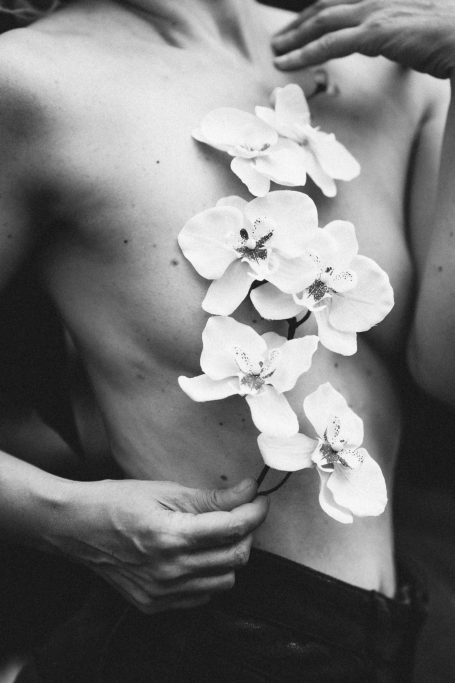Detailaufnahme eines weiblichen Körpers im Teilakt mit weißen Orchideen auf der Haut und Händen, die die Blüten halten.