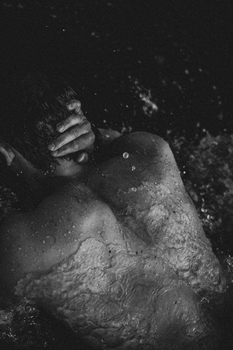 Künstlerische Schwarz-Weiß-Aktaufnahme eines Männerrückens im Wasser, während Wellen und Wassertropfen über den Körper
laufen