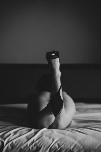 Schwarz-Weiß-Aufnahme einer auf dem Bett liegenden Frau mit angewinkelten Beinen und schwarzen High Heels.