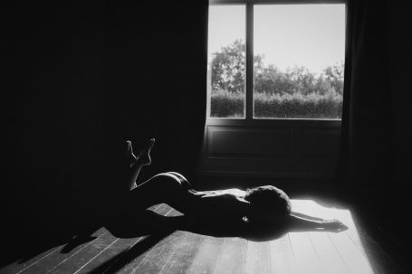 Teilakt einer Frau, die im starken Sonnenlicht auf einem Holzboden liegt. Licht und Schatten zeichnen die Silhouette ihres Körpers nach.