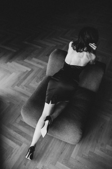 Frau in einem schwarzen Kleid und High Heels liegt elegant auf einem Sessel, aus erhöhter Perspektive in Schwarz-Weiß fotografiert.