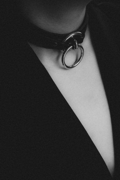 Künstlerische Schwarz-Weiß-Detailaufnahme eines schwarzen Halsbands mit großem Metallring am Hals einer Frau.