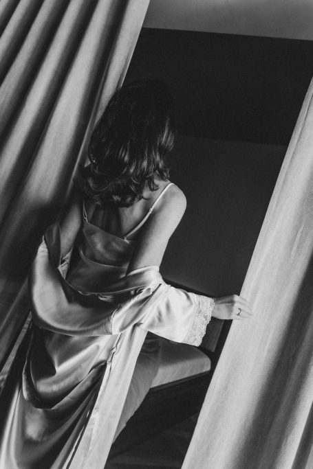 Frau in einem Satinmorgenmantel zwischen schweren Vorhängen, von hinten in stimmungsvollem Schwarz-Weiß fotografiert.