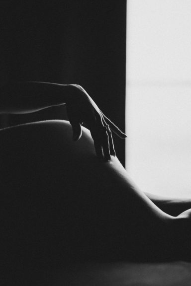 Schwarz-Weiß-Detailaufnahme einer Hand auf dem Körper einer Frau mit starkem Licht-und-Schatten-Kontrast am Fenster.