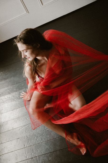 Frau in roter Lingerie sitzt auf dem Boden, während transparenter roter Tüll ihren Körper weich umspielt.
