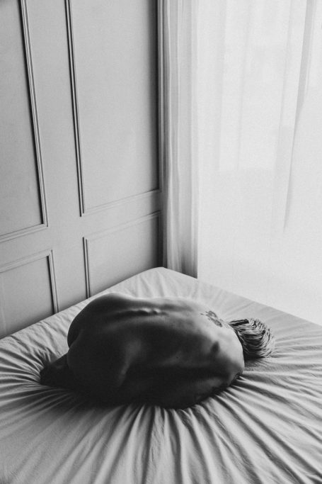 Unbekleidete Frau in zusammengezogener Pose auf einem Bett, fotografiert in kontrastreichem Schwarz-Weiß