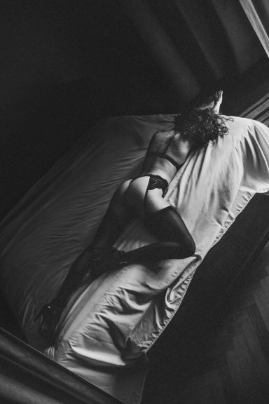 Schwarz-Weiß-Aufnahme einer Frau in schwarzer Lingerie und Strümpfen, die ausgestreckt auf einem Bett liegt.