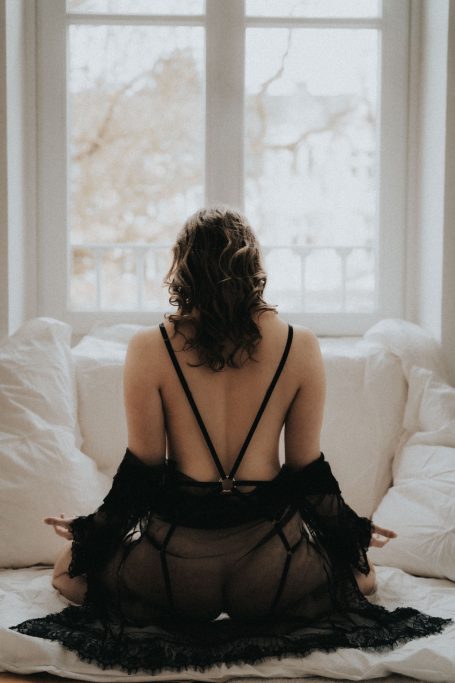 Rückansicht einer Frau in schwarzer Lingerie und Spitzenrobe auf einem Bett vor einem großen Fenster.