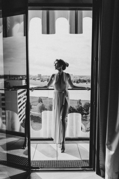 Schwarz-Weiß-Porträt einer Frau in einem langen fließenden Kleid vor einer geöffneten Balkontür mit weitem Ausblick.