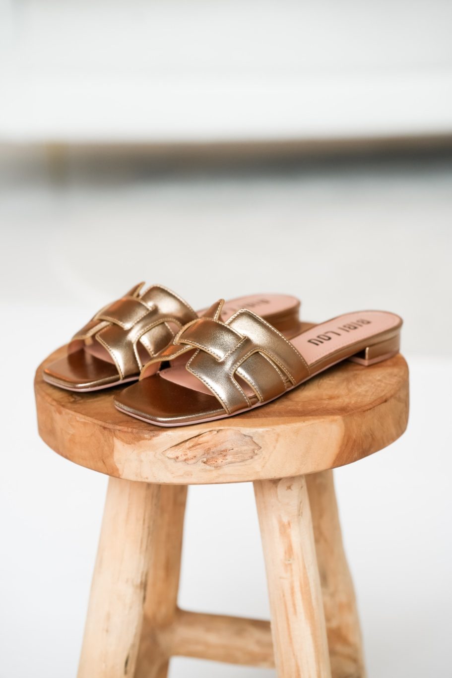 Goldfarbene flache Sandalen mit geflochtenem Riemendetail auf einem runden Holzhocker.