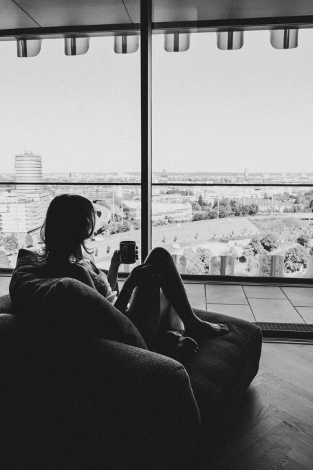 Schwarz-Weiß-Aufnahme einer Frau, die mit einer Tasse im Sessel vor einem großen Fenster mit Stadtblick sitzt.